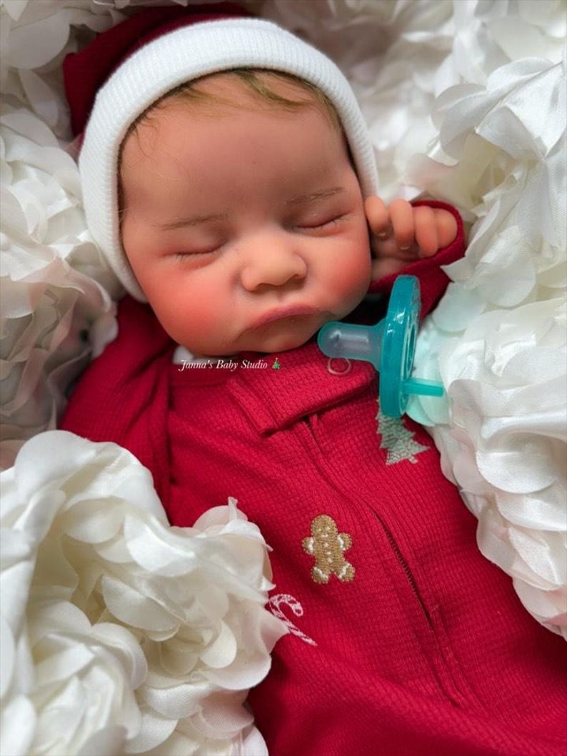 May include: A lifelike baby doll wearing a red onesie with a gingerbread man and Christmas tree applique. The doll has a white beanie and a blue pacifier. The doll is nestled in white fabric flowers. The text "Jamma's Baby Studio" is visible.