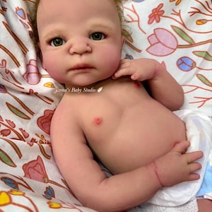 May include: A lifelike baby doll with fair skin, green eyes, and reddish-brown hair. The doll is lying on a floral patterned blanket and wearing a diaper. The doll's chest has two small red marks. The text "Janna's Baby Studio" is visible.