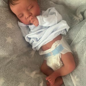 May include: A realistic baby doll is lying on a soft, gray blanket with a star pattern. The doll is wearing a light blue onesie and a diaper. The baby is sleeping with its hand near its mouth. The text "Lanna's Baby Studio" is visible.