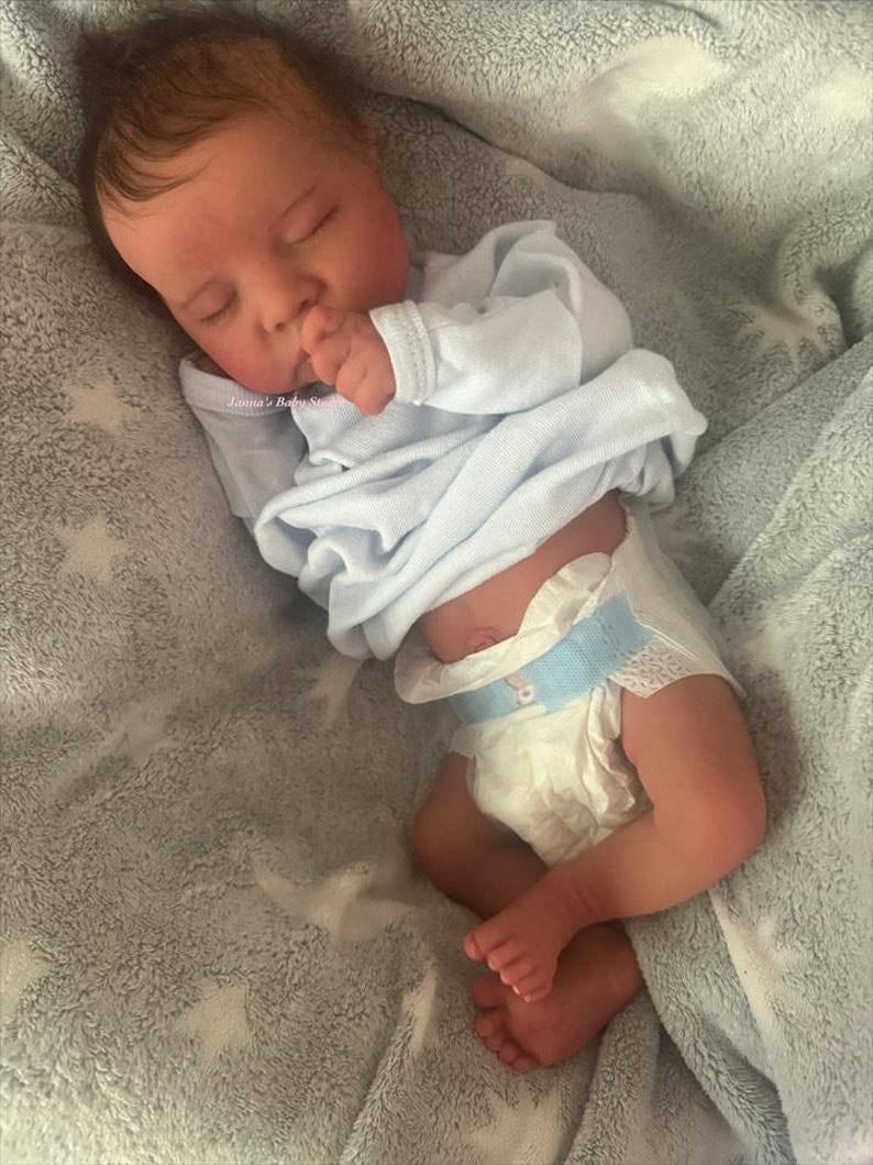 May include: A realistic baby doll is lying on a soft, gray blanket with a star pattern. The doll is wearing a light blue onesie and a diaper. The baby is sleeping with its hand near its mouth. The text "Lanna's Baby Studio" is visible.