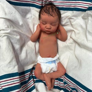 May include: A lifelike reborn baby doll with closed eyes, lying on a white and blue striped blanket. The doll has a realistic skin tone, a diaper, and a small tuft of dark hair. The doll's arms are raised near its face.