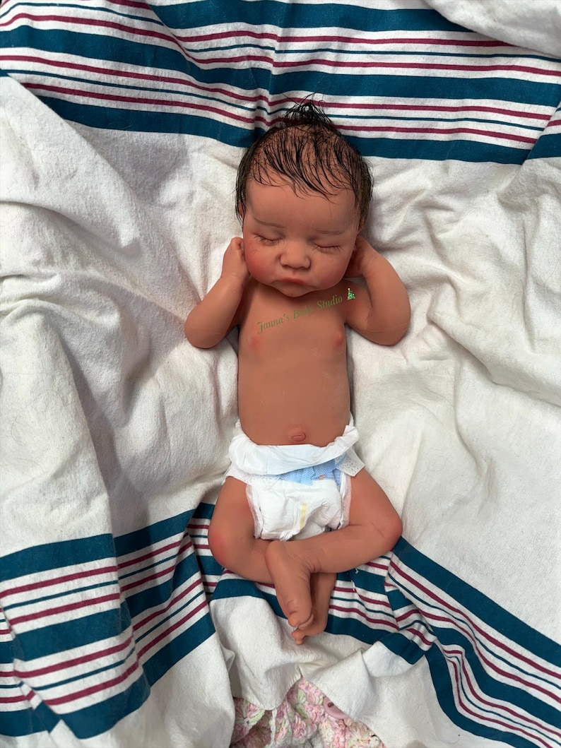 May include: A lifelike reborn baby doll with closed eyes, lying on a white and blue striped blanket. The doll has a realistic skin tone, a diaper, and a small tuft of dark hair. The doll's arms are raised near its face.