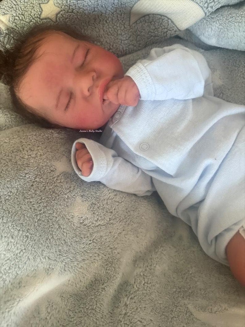 May include: A baby doll wearing a light blue long-sleeved onesie. The doll is lying on a soft, light blue blanket with a star pattern.