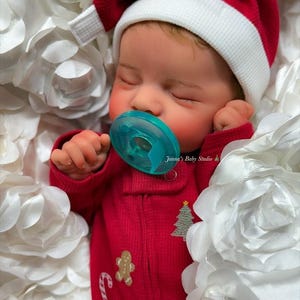 May include: A sleeping baby wearing a red Santa hat with a white brim and a red onesie. The onesie has a gingerbread man, candy cane, and Christmas tree design. The baby is using a blue pacifier and is surrounded by white fabric flowers. The text "Janna's Baby Studio" is visible.