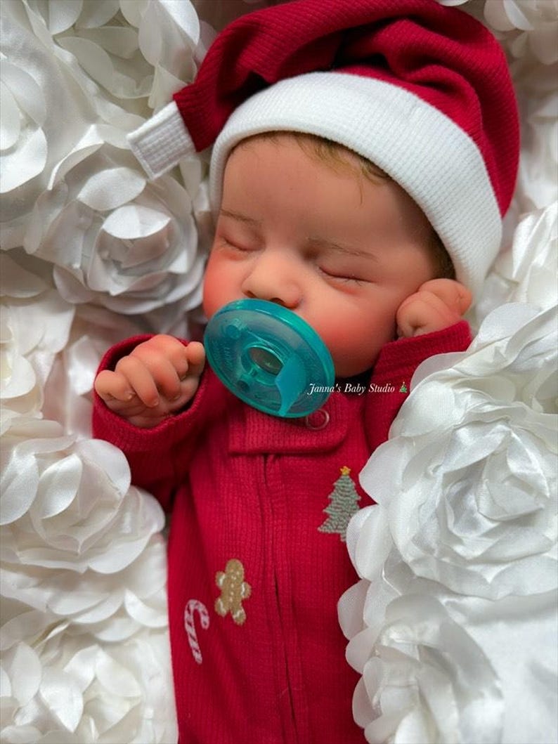 May include: A sleeping baby wearing a red Santa hat with a white brim and a red onesie. The onesie has a gingerbread man, candy cane, and Christmas tree design. The baby is using a blue pacifier and is surrounded by white fabric flowers. The text "Janna's Baby Studio" is visible.