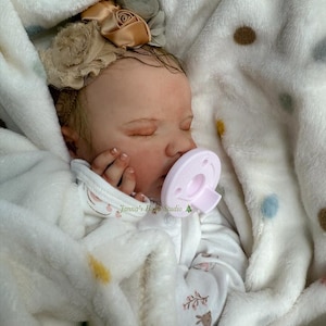 May include: A realistic baby doll asleep, swaddled in a white, fluffy blanket with a polka dot design. The doll wears a white onesie with a floral pattern and a beige floral headband. A pink dummy is in its mouth. The text "Janna's Baby Studio" is visible.