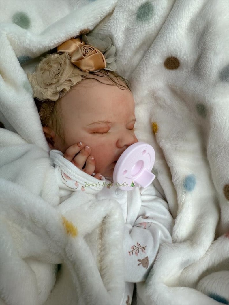 May include: A realistic baby doll asleep, swaddled in a white, fluffy blanket with a polka dot design. The doll wears a white onesie with a floral pattern and a beige floral headband. A pink dummy is in its mouth. The text "Janna's Baby Studio" is visible.