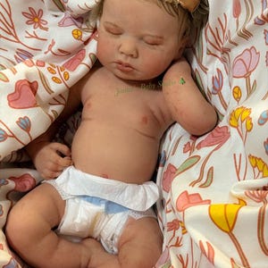 May include: A realistic baby doll with closed eyes, wearing a nappy and a floral headband. The doll is lying in a floral patterned blanket. The text "Janna Baby Studio" is visible on the doll's arm.