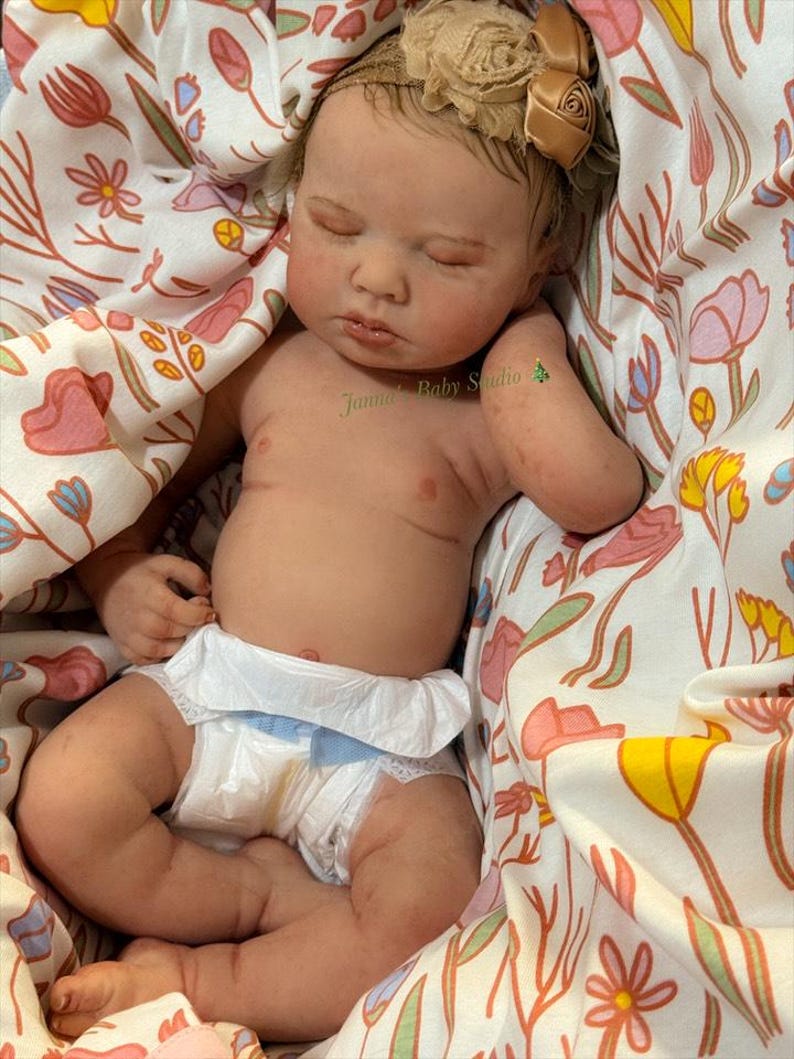May include: A realistic baby doll with closed eyes, wearing a nappy and a floral headband. The doll is lying in a floral patterned blanket. The text "Janna Baby Studio" is visible on the doll's arm.