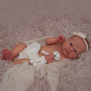 May include: A realistic baby doll with a white crocheted blanket and a white crocheted stuffed animal. The doll is wearing a white diaper and a headband with a white flower.
