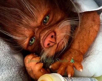 May include: A realistic doll with reddish-brown skin, green eyes, and dark hair covering its face and arms. The doll has a snout-like nose and is holding a plastic baby bottle with a yellow nipple and a label reading Enfamil Enfacare.