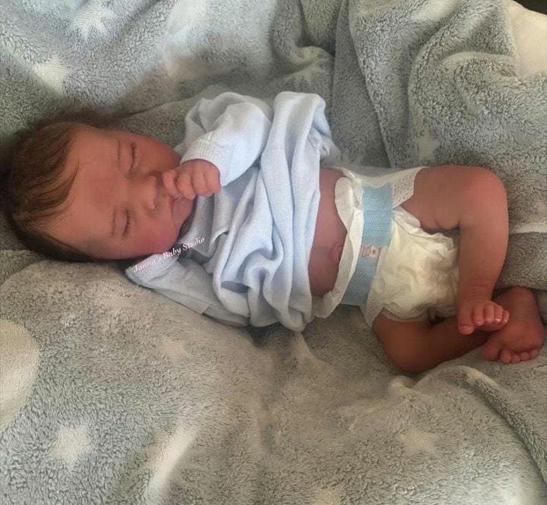 May include: A realistic baby doll is lying on a soft, gray blanket. The doll is wearing a light blue onesie and a diaper. The baby's eyes are closed, and it appears to be sleeping. The doll's skin has a realistic tone.