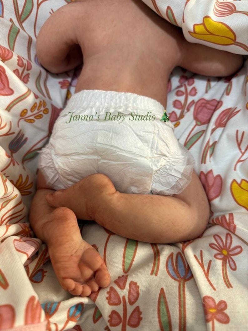 May include: A baby doll, lying prone, wearing a white nappy. The doll is on a floral patterned blanket. The text "Janna's Baby Studio" is visible.