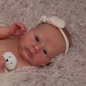 May include: A realistic reborn baby doll with brown eyes, a light pink complexion, and a white headband with a pearl accent. The doll is lying on a white fluffy blanket.