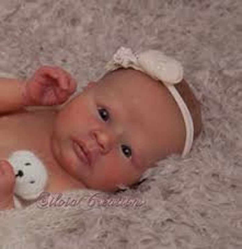 May include: A realistic reborn baby doll with brown eyes, a light pink complexion, and a white headband with a pearl accent. The doll is lying on a white fluffy blanket.