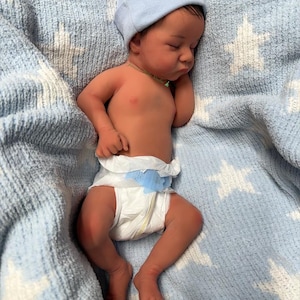 May include: A newborn baby wearing a light blue beanie and a diaper, sleeping peacefully on a blue blanket with white star patterns. The baby has dark hair and a small green necklace.