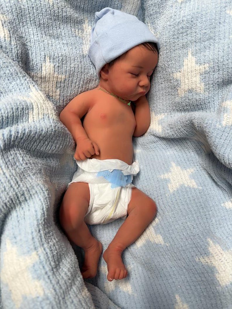 May include: A newborn baby wearing a light blue beanie and a diaper, sleeping peacefully on a blue blanket with white star patterns. The baby has dark hair and a small green necklace.