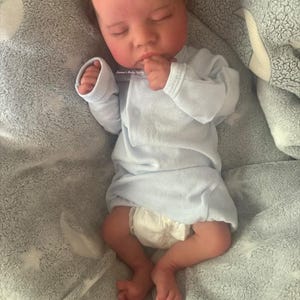 May include: A realistic baby doll is lying on a gray blanket. The doll is wearing a light blue onesie and a diaper. The baby has closed eyes and is resting its hand near its mouth. The blanket has a star pattern.