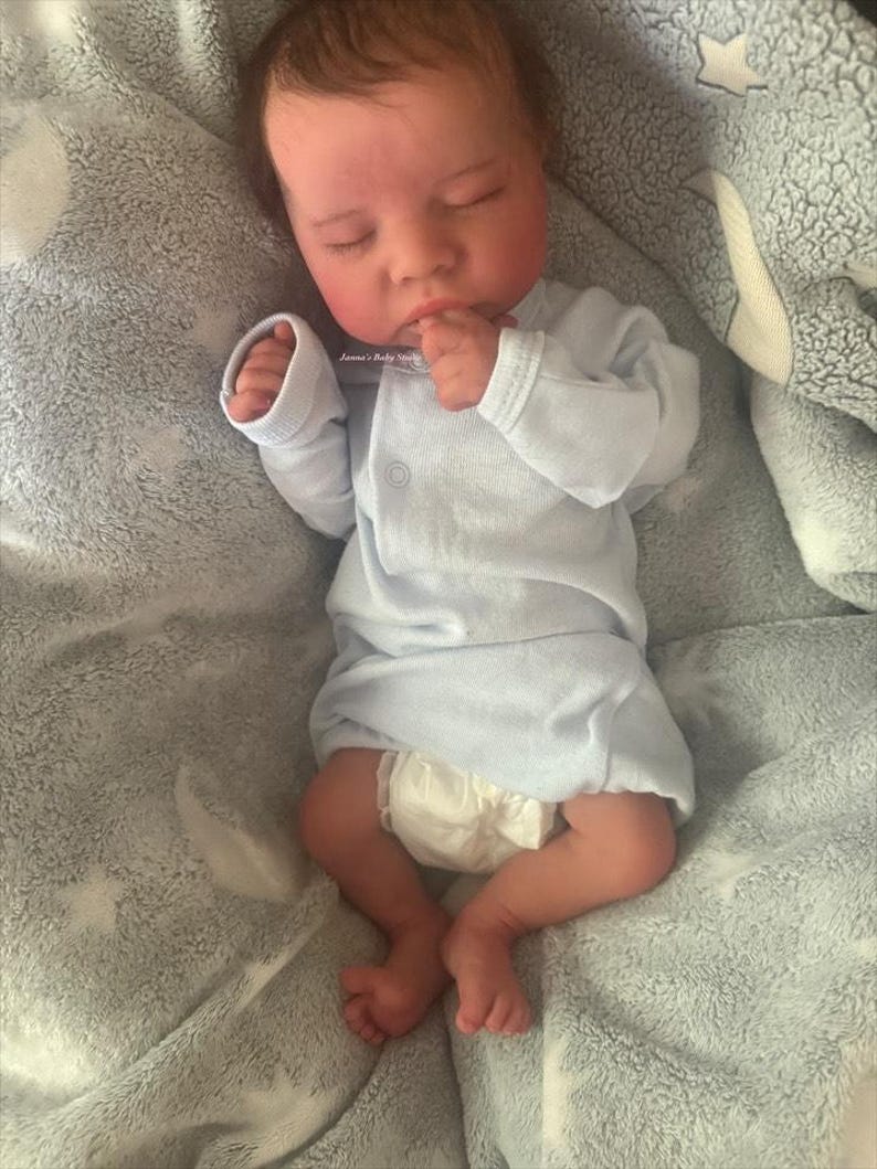 May include: A realistic baby doll is lying on a gray blanket. The doll is wearing a light blue onesie and a diaper. The baby has closed eyes and is resting its hand near its mouth. The blanket has a star pattern.