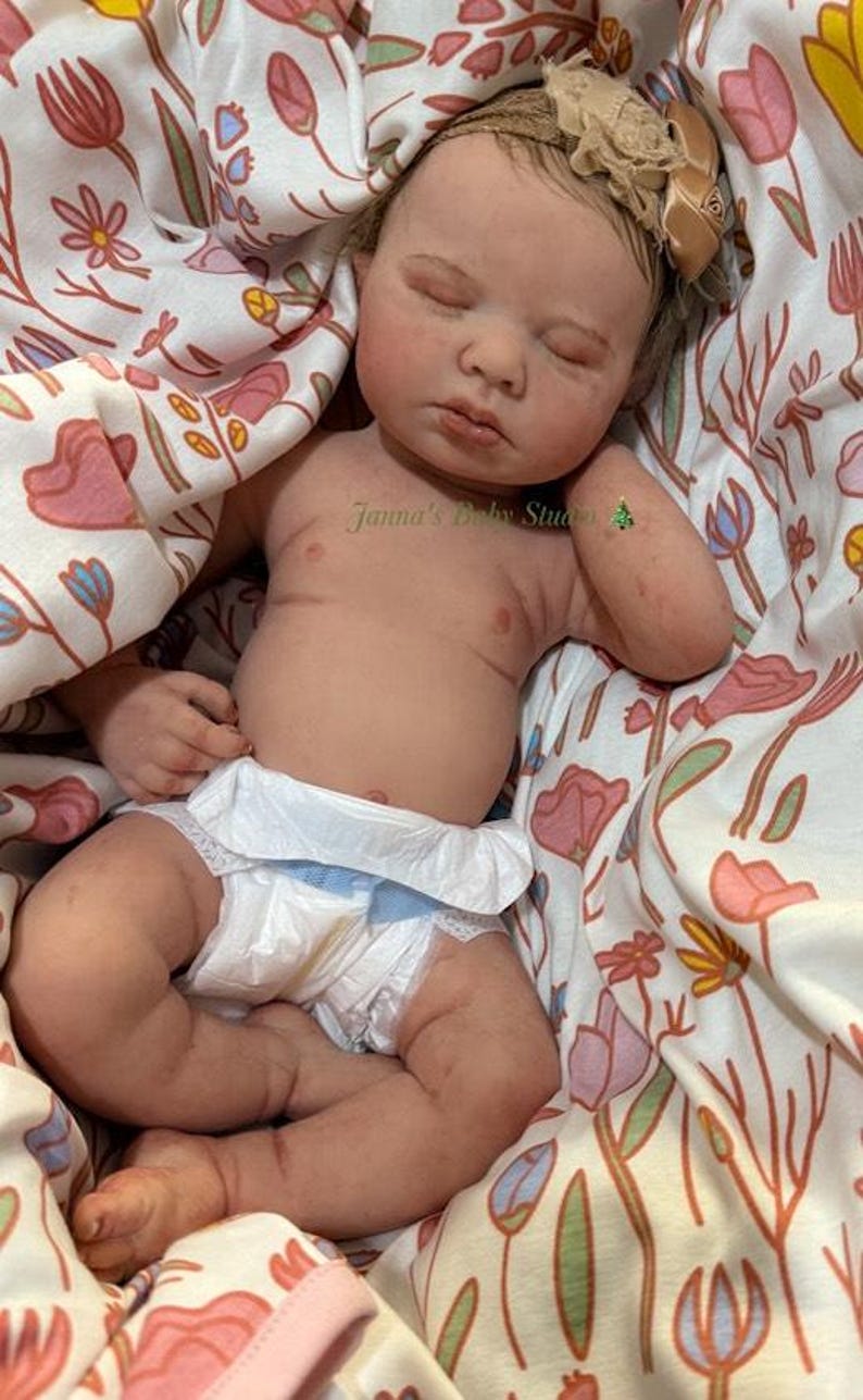 May include: A realistic baby doll with closed eyes, wearing a nappy, and a floral headband. The doll is lying on a floral patterned blanket. The text "Janna's Baby Studio" is visible on the doll's body.