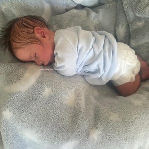 May include: A sleeping baby wearing a light blue onesie and a diaper, resting on a soft, gray blanket with a star pattern. The baby has dark hair and is curled up on the blanket.