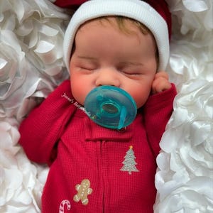 May include: A sleeping baby doll wearing a red onesie with a Christmas tree, gingerbread man, and candy cane design. The doll is wearing a white and red Santa hat and has a blue pacifier. The doll is nestled in white fabric.