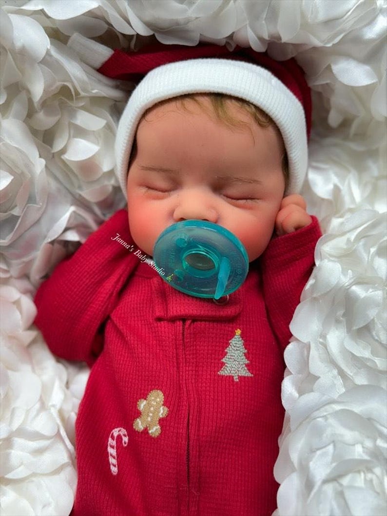 May include: A sleeping baby doll wearing a red onesie with a Christmas tree, gingerbread man, and candy cane design. The doll is wearing a white and red Santa hat and has a blue pacifier. The doll is nestled in white fabric.