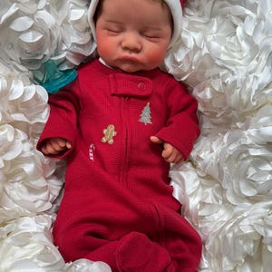 May include: A sleeping baby wearing a red Christmas-themed onesie with a Santa hat. The onesie features a gingerbread man, a Christmas tree, and a candy cane. The baby is nestled in a bed of white, flower-shaped fabric.