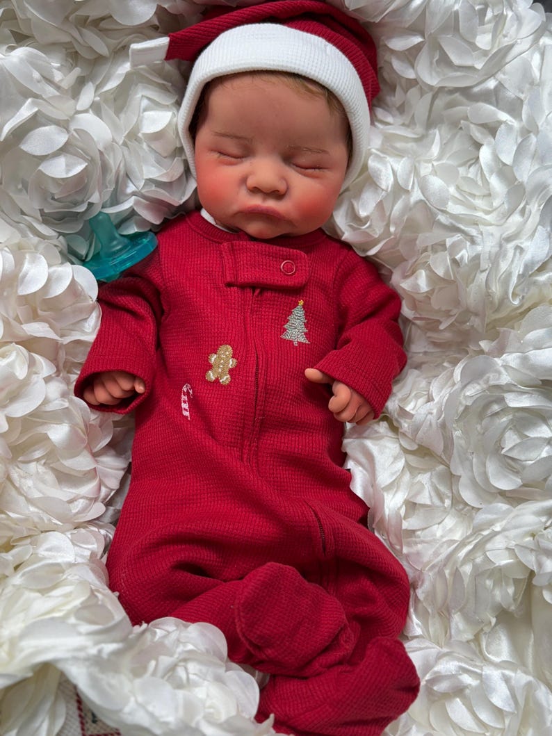 May include: A sleeping baby wearing a red Christmas-themed onesie with a Santa hat. The onesie features a gingerbread man, a Christmas tree, and a candy cane. The baby is nestled in a bed of white, flower-shaped fabric.
