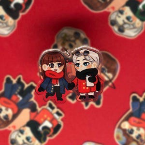 May include: Two small, cartoon figures of a couple, one in a red coat and the other in a blue coat, with red scarves, stand on a red background with a repeating pattern of the same figures. The figures are holding hands.