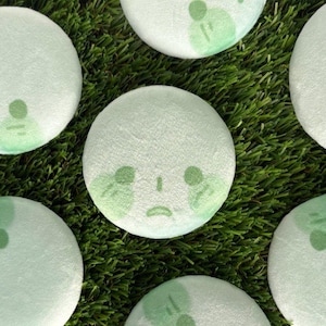 Peut inclure: Plusieurs coussins ronds vert clair avec un motif de visage triste en vert plus foncé. Ils sont disposés sur de l'herbe artificielle verte. Les coussins ont un aspect doux et texturé.