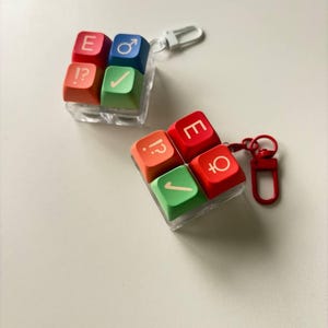 May include: Two square keycap keychains featuring four colorful keys each. One keychain has red, blue, orange, and green keys with symbols E, male sign, !?, and a checkmark. The other has red, orange, and green keys with E, !?, and female sign.