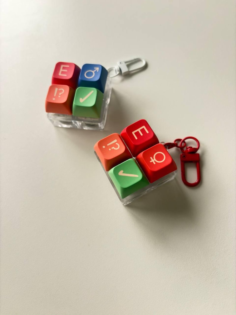 May include: Two square keycap keychains featuring four colorful keys each. One keychain has red, blue, orange, and green keys with symbols E, male sign, !?, and a checkmark. The other has red, orange, and green keys with E, !?, and female sign.