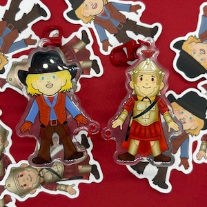 以下が含まれることがあります： 赤い背景に、漫画のキャラクターのキーホルダーとステッカーのコレクション。キーホルダーはカウボーイとローマ兵士を特徴としています。ステッカーは同じキャラクターをさまざまなポーズで描いています。カウボーイのキーホルダーは黒い帽子とオレンジ色のベストを持っています。