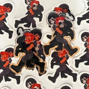 Puede incluir: Una colección de pegatinas y llaveros con un personaje de dibujos animados con pelo rojo y una máscara de calavera, que lleva a otro personaje. Los personajes visten ropa oscura con detalles dorados. El fondo es blanco.