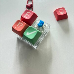 May include: A square clear plastic keychain featuring three mechanical keyboard keycaps in red, orange, and green, with white symbols including an E, a checkmark, and an interrobang. A separate red keycap with a female gender symbol sits nearby.