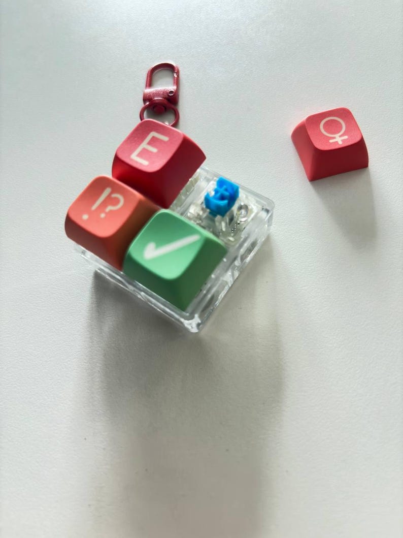 May include: A square clear plastic keychain featuring three mechanical keyboard keycaps in red, orange, and green, with white symbols including an E, a checkmark, and an interrobang. A separate red keycap with a female gender symbol sits nearby.