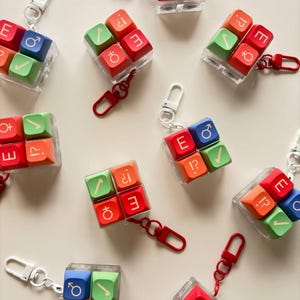 May include: A collection of square keycap keychains, each featuring four colorful keys arranged in a grid. The keys are red, green, blue, and orange, displaying various symbols including gender signs, checkmarks, and punctuation marks.