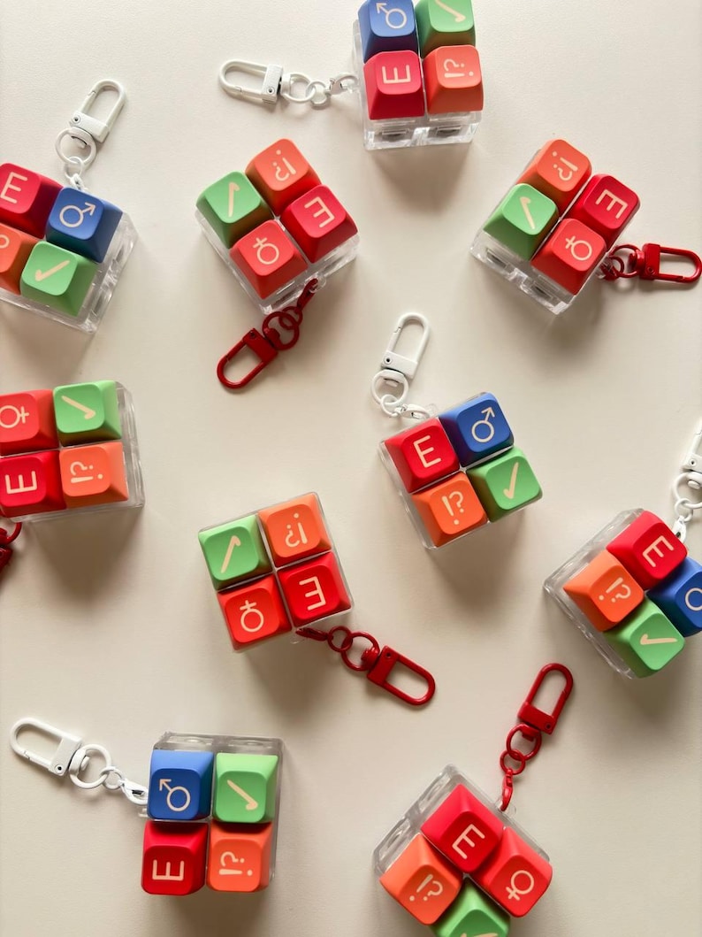 May include: A collection of square keycap keychains, each featuring four colorful keys arranged in a grid. The keys are red, green, blue, and orange, displaying various symbols including gender signs, checkmarks, and punctuation marks.