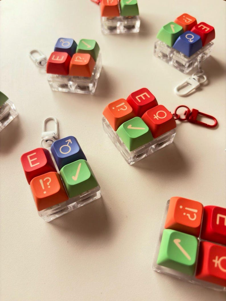 May include: Several square keycap keychains featuring four colorful mechanical keyboard keys arranged in a grid. Each keycap displays symbols including the letter E, a checkmark, a question mark with an exclamation point, and gender symbols.