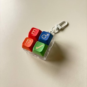 May include: A square keychain featuring four colorful keyboard keycaps arranged in a grid. The top-left key is red with the letter E, the top-right is blue with a male symbol, the bottom-left is orange with !?, and the bottom-right is green with a checkmark.