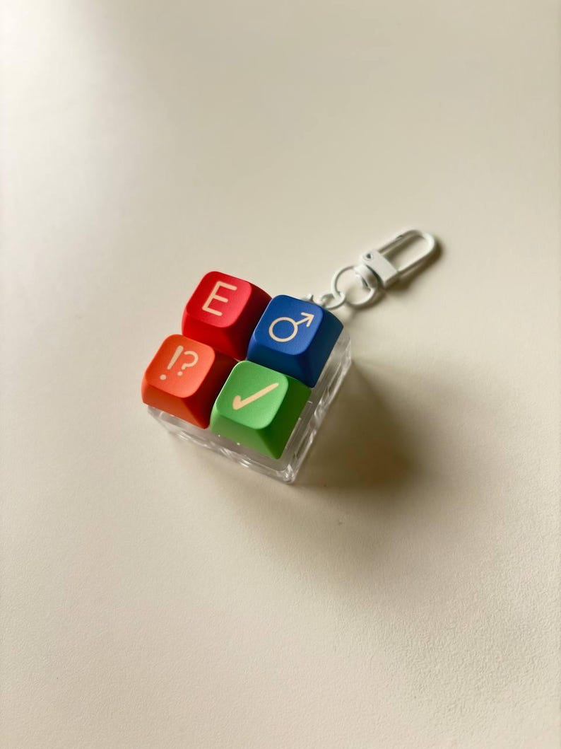May include: A square keychain featuring four colorful keyboard keycaps arranged in a grid. The top-left key is red with the letter E, the top-right is blue with a male symbol, the bottom-left is orange with !?, and the bottom-right is green with a checkmark.