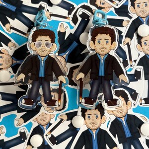 Puede incluir: Dos llaveros acrílicos transparentes con una ilustración de dibujos animados de un hombre con traje, camisa azul y gafas. Los llaveros tienen un cierre azul. El fondo es una superficie azul con ilustraciones similares y pastillas blancas.