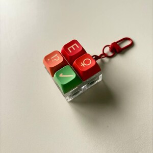 May include: A square keychain featuring four mechanical keyboard keycaps arranged in a two-by-two grid. The top left keycap is orange with a question mark and period, the top right is red with an E, the bottom left is green with a checkmark, and the bottom right is red with a female gender symbol. A red metal clasp is attached.
