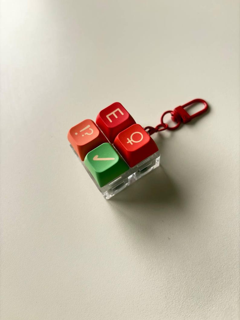 May include: A square keychain featuring four mechanical keyboard keycaps arranged in a two-by-two grid. The top left keycap is orange with a question mark and period, the top right is red with an E, the bottom left is green with a checkmark, and the bottom right is red with a female gender symbol. A red metal clasp is attached.