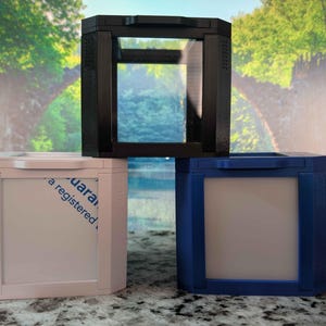 May include: Three geometric boxes in black, pink, and blue. Each box has a rectangular opening and a white interior. The black box is positioned above the other two. The background features a scenic landscape with trees and water.