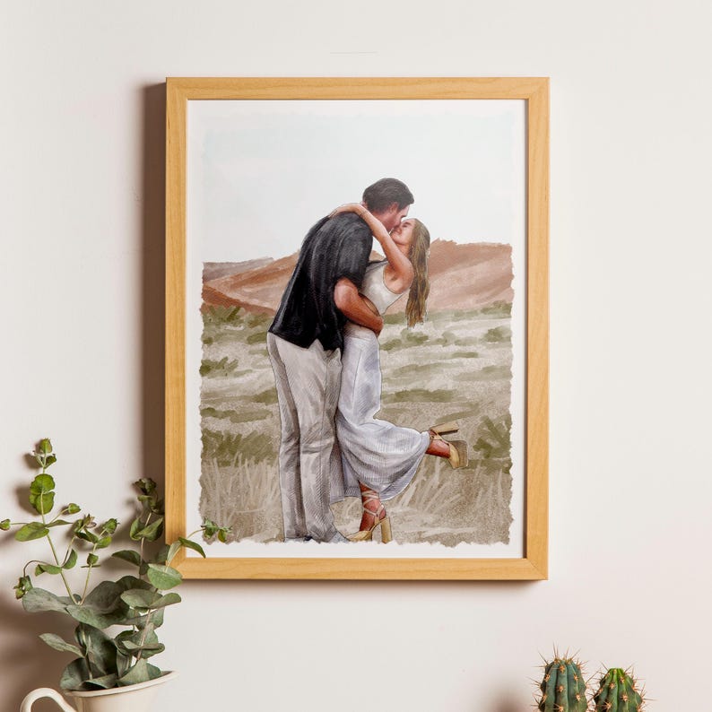 May include: A framed watercolour illustration depicting two people embracing and kissing in a desert landscape. One person wears a black shirt and light trousers, while the other wears a white dress and tan heels. The artwork is in a light wood frame.