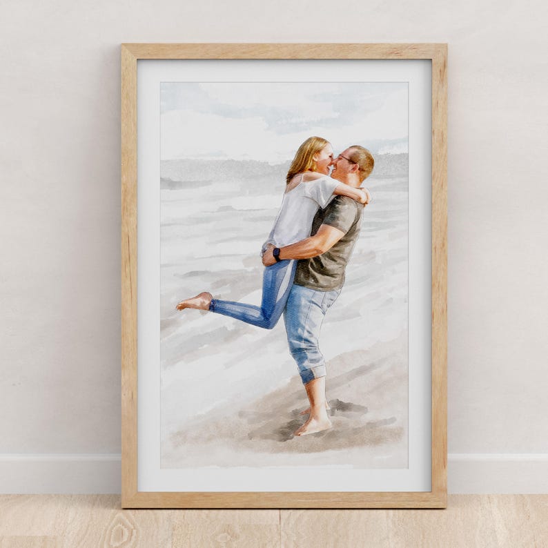 May include: A watercolour-style art print in a light wood frame depicting a person lifting another person in their arms on a beach. The person being lifted wears a white top and blue jeans, while the other wears a dark top and blue jeans.