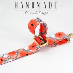 May include: A close-up of a red and white floral printed strap with a gold metal buckle. The strap is handmade and is labeled "HANDMADE Printed Straps".