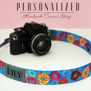 Può includere: Un cinturino per fotocamera blu con un motivo a ciambelle e il nome "LILY" stampato su una toppa grigia. Il cinturino è attaccato a una fotocamera nera.